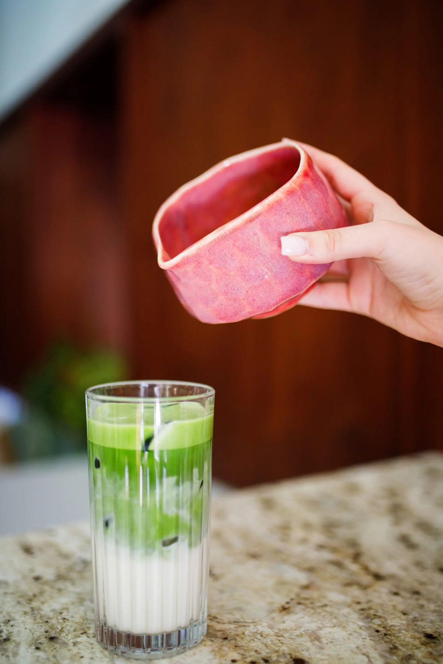 Hand holding a pink ceramic container over a glass with a layered drink on a countertop.