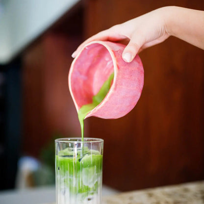 Person pouring a green liquid from a pink container into a glass on a countertop.