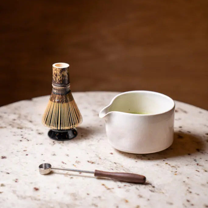 Tea-making tools including a whisk, chasaku, and chawan on a marble surface with a dark background.