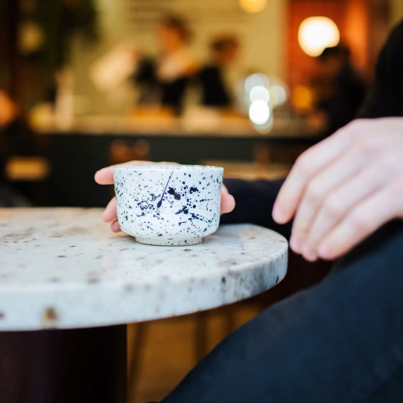 Person holding a speckled ceramic cup on a marble table with a blurred cafe background