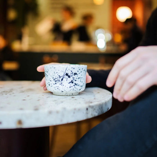 Person holding a speckled ceramic cup on a marble table with a blurred cafe background
