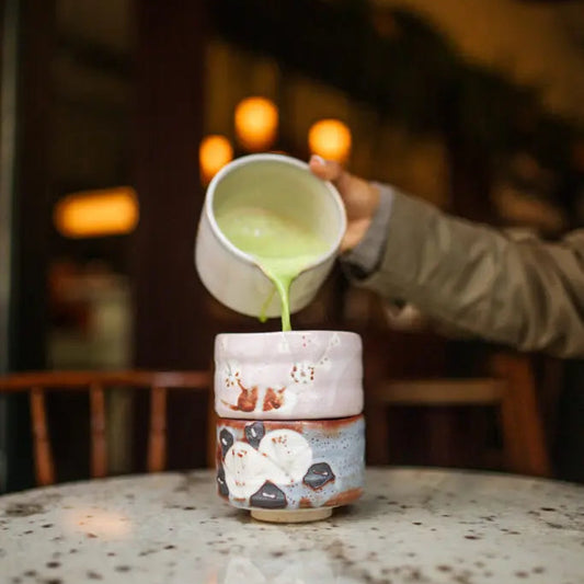 Person pouring green liquid from a small cup into a larger, decorated cup on a table.