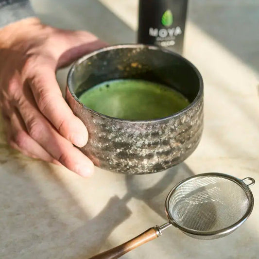 Person holding a textured ceramic cup with green liquid, next to a Moya coffee can and a metal sieve on a light surface.