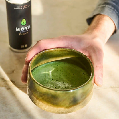 Hand holding a green ceramic bowl with a can labeled 'Moya' in the background.