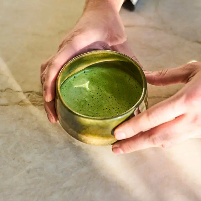 Person holding a green ceramic cup filled with a green liquid on a textured surface