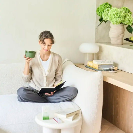 Person sitting on a white couch holding a green mug and a book in a modern living room.
