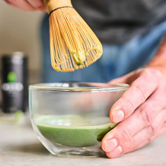 Person using a wooden whisk to prepare matcha green tea in a glass bowl.